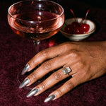 Esme ring on a hand beside a coupe glass at a night-time bar