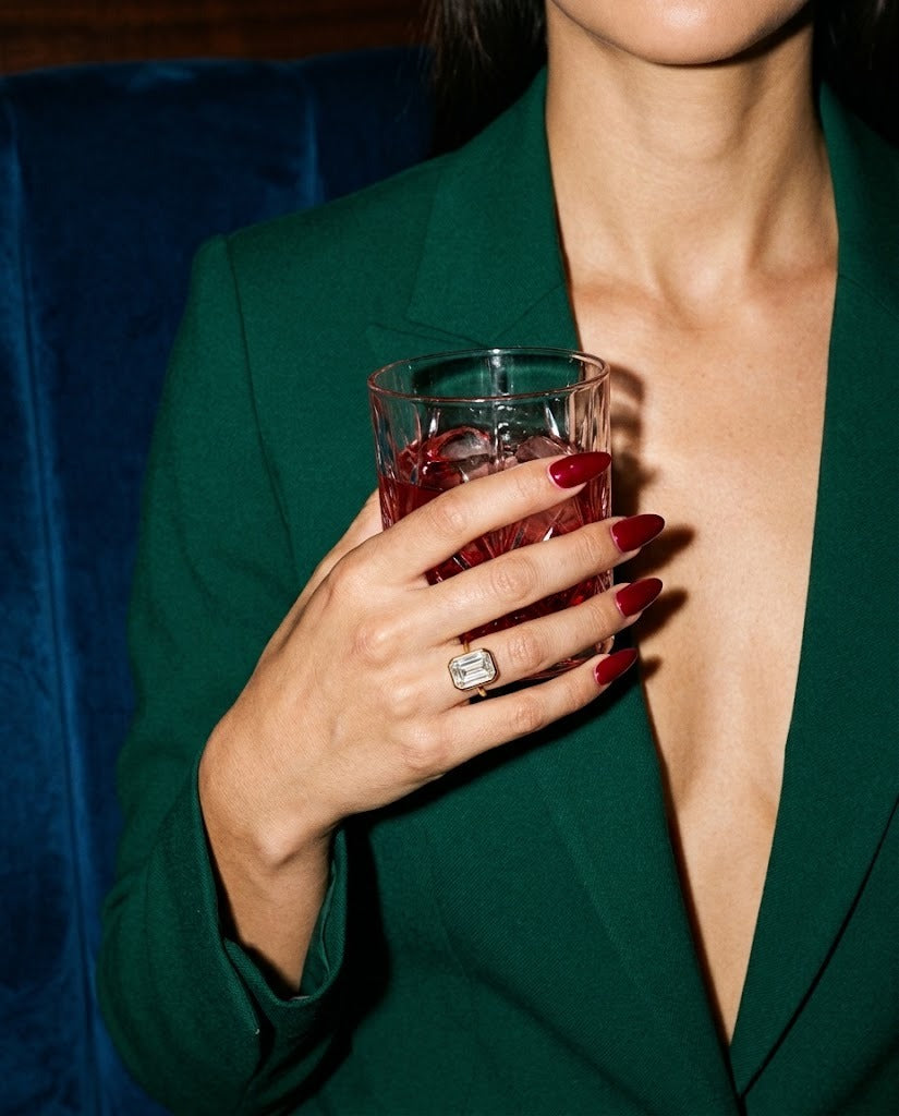 Esme ring on a hand holding a crystal tumbler, emerald suit, night-time
